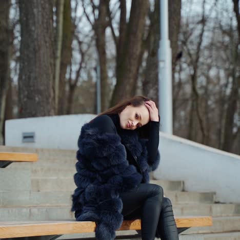 luxury girl sitting on bench. attractive young woman in fur coat enjoys fresh air in the park and smiles. positive brunette outdoors.