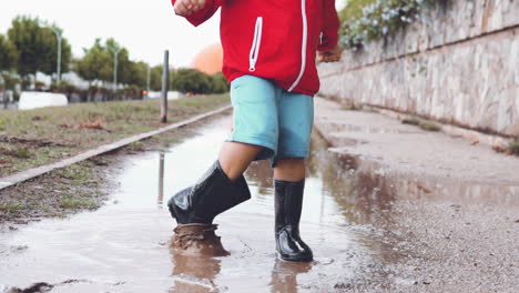 little boy wearing rubber boots splashing water from a puddle to the camera in slow motion