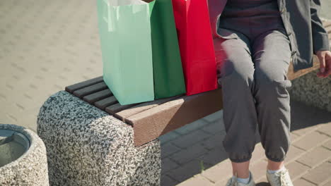 close-up leg view of a woman wearing grey joggers as she drops her shopping bags on a wooden bench, she sits down and crosses her legs while wearing white sneaker