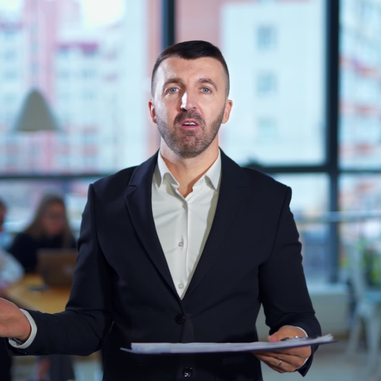 Serious man in suit talks in front of camera showing graphs. Confident male speaker performs presentation in office. People work at the desk at backdrop in blur