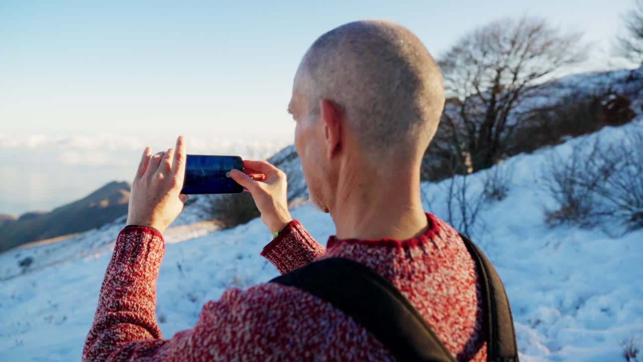 Rear view of a man using a smartphone to capture a snowy mountain landscape