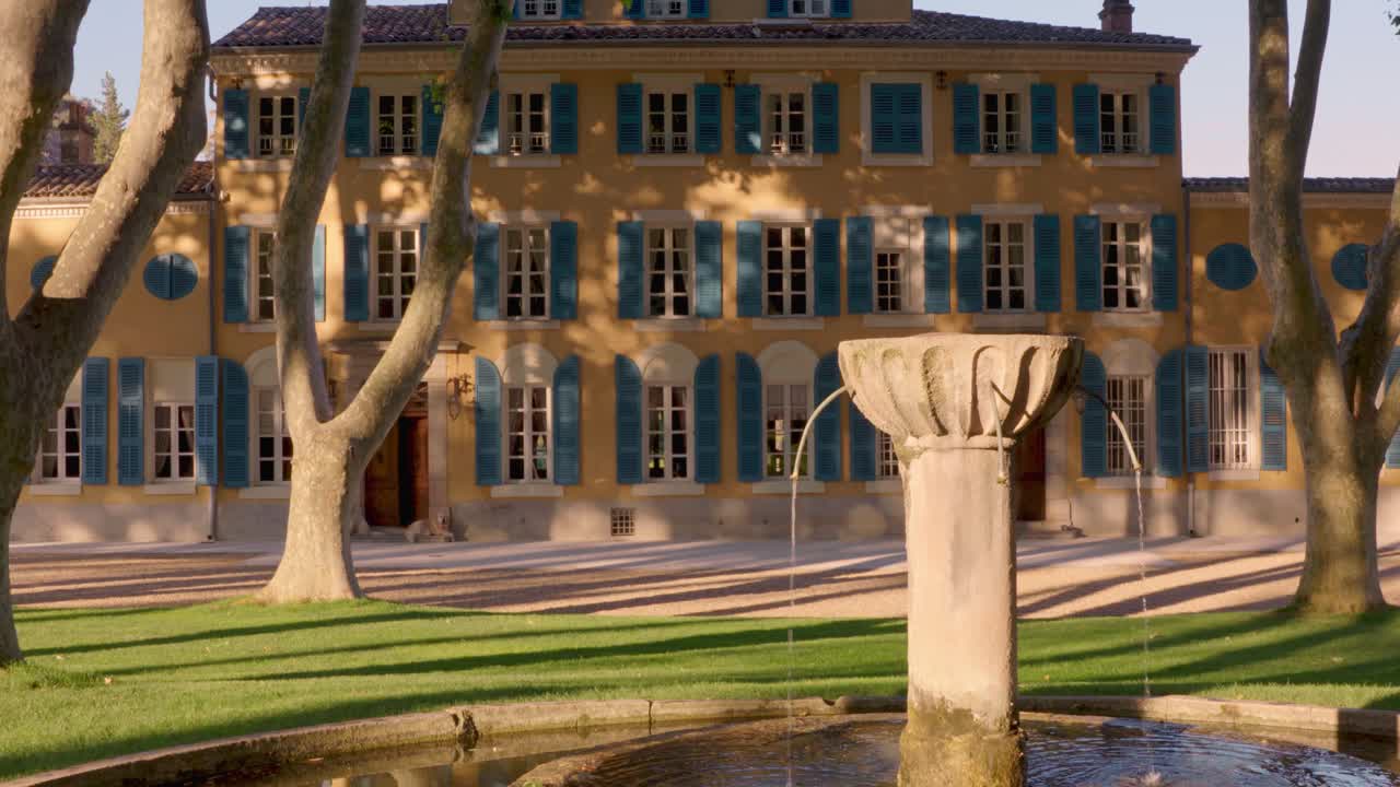 A wide shot captures a grand yellow mansion with blue shutters, framed by trees and a stone fountain in the foreground, set in a serene garden.