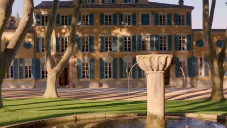 a wide shot captures a grand yellow mansion with blue shutters, framed by trees and a stone fountain in the foreground, set in a serene garden.
