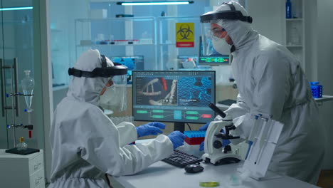 Chemist-in-ppe-suit-working-at-pc-while-lab-technician-bringing-her-blood-samples