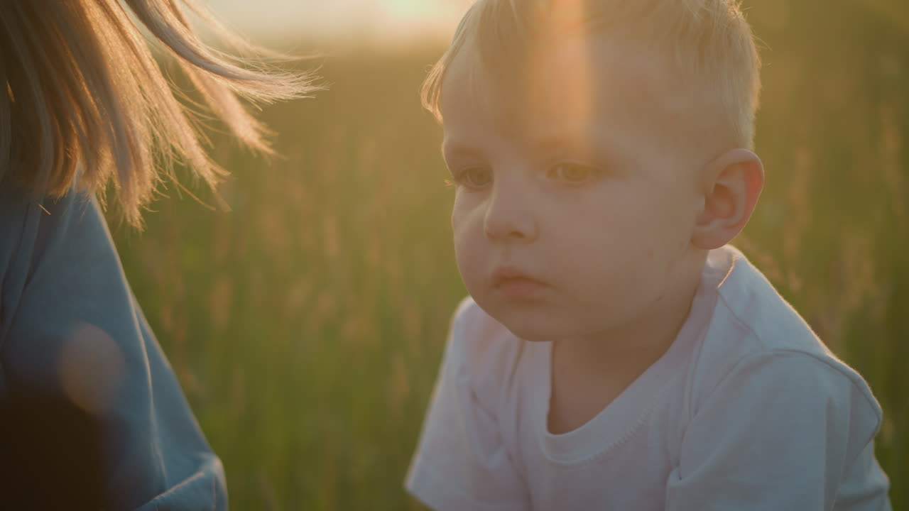Close-up of a young boy in a white shirt, looking contemplative in a grassy field at sunset, with a woman partially visible in the background