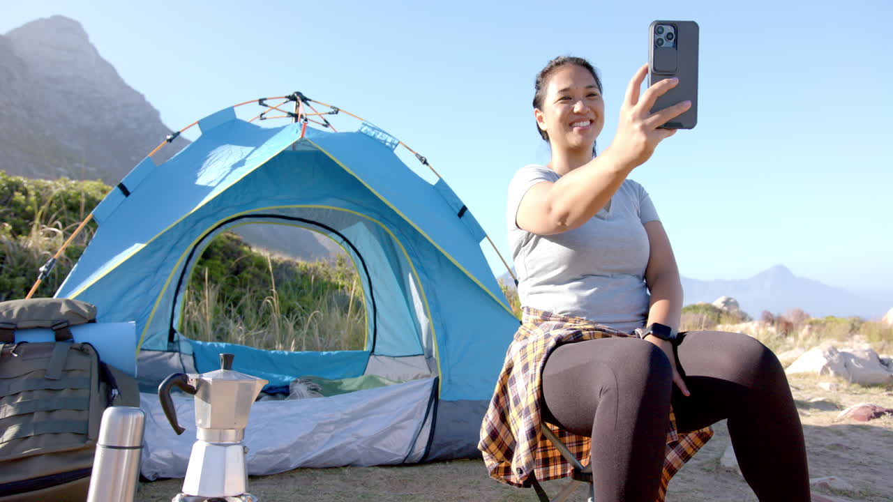 Taking selfie, woman sitting by tent during mountain hike, smiling outdoors