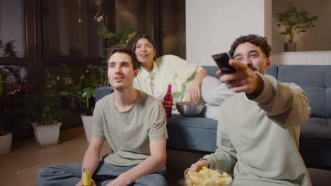 three friends watching comic movie on television sitting on couch and floor, eating chips and drinking soda