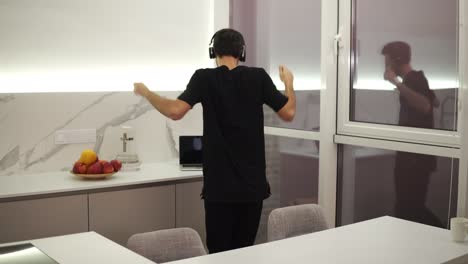 Joyful-man-in-black-clothes-is-dancing-in-kitchen-listening-to-music-with-headphones,-having-fun-alone-in-light-modern-apartment.-Devices-and-people-concept.-Accelerated