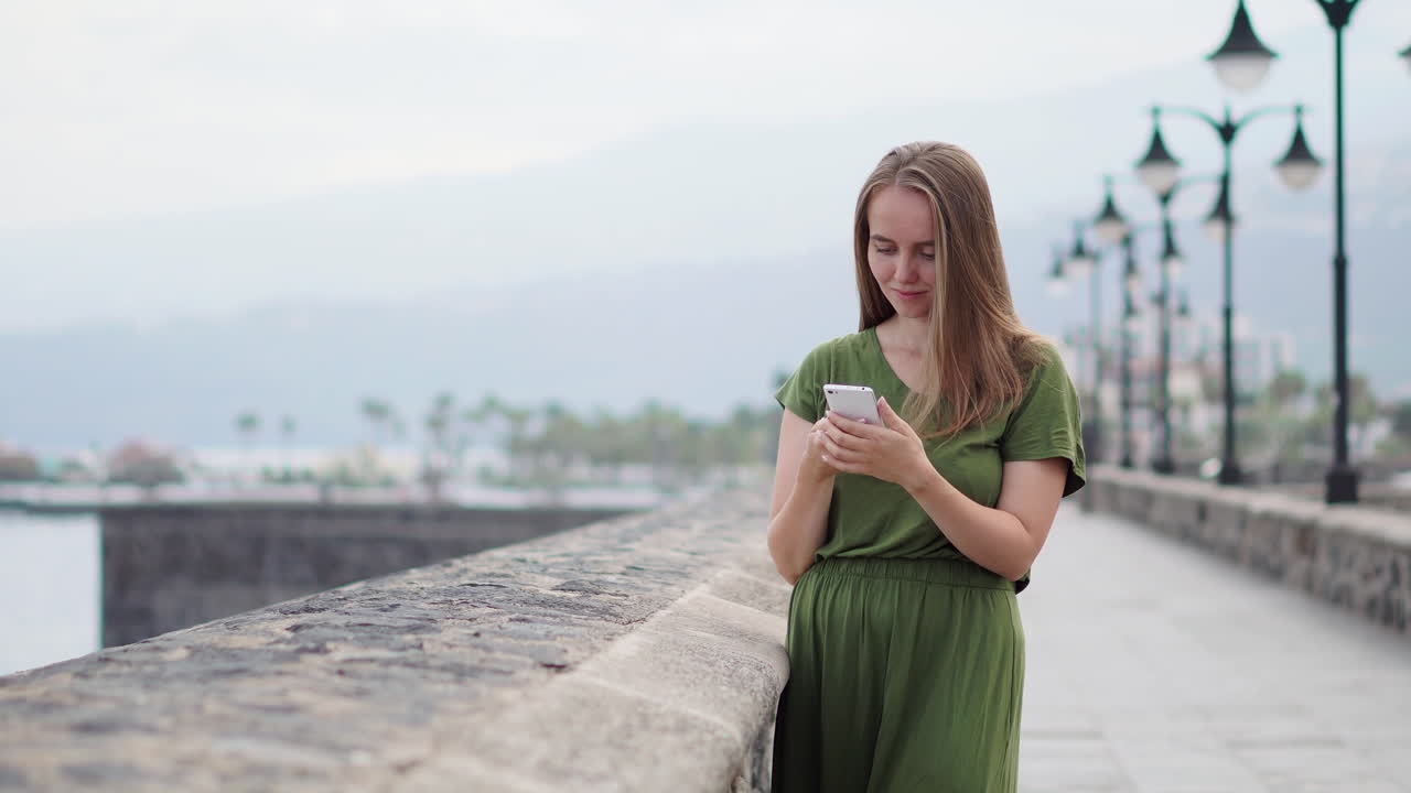 With the seascape horizon of a beach in view, a traveler operates a mobile phone in her hand. The tourist's attention is drawn to the ocean's tranquil blue expanse and the sun's warmth, reflecting a lively summer lifestyle