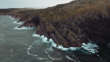 aerial view from drone flying toward rocky shoreline with waves breaking against cliffs