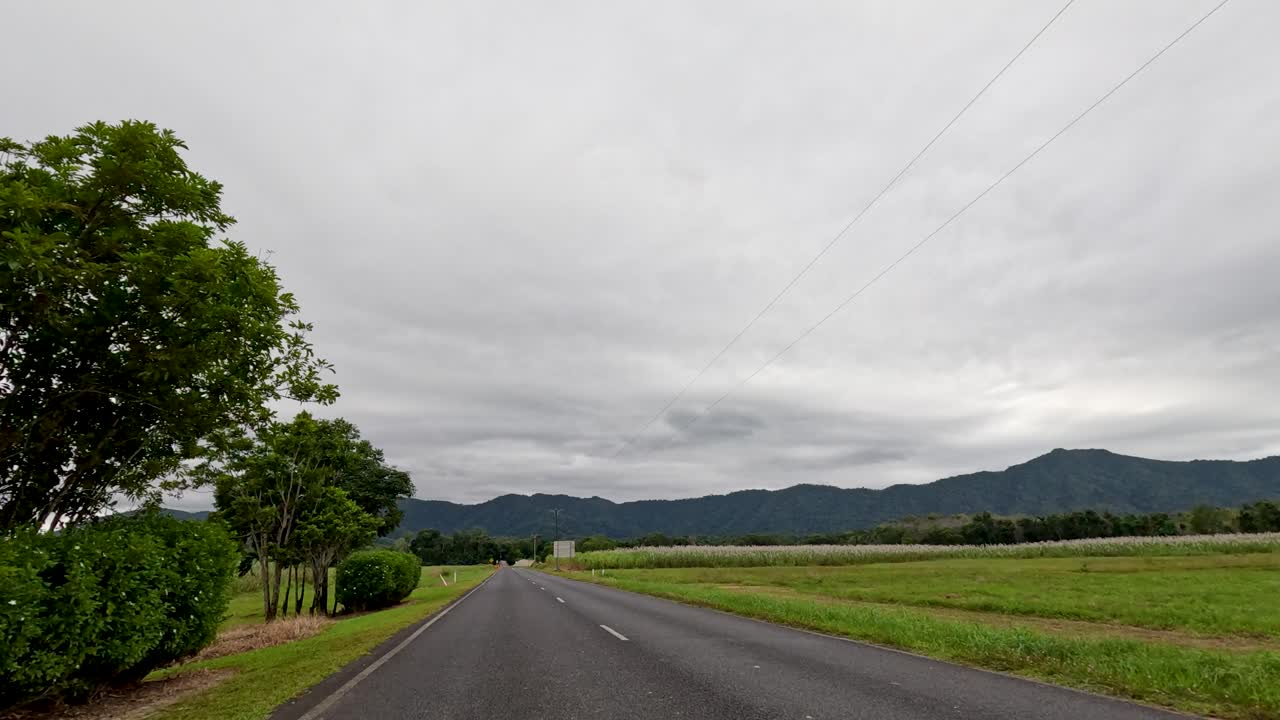 A serene drive along a rural road in Port Douglas, showcasing lush greenery and distant mountains under overcast skies