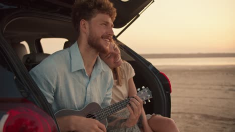 a bearded man with curly hair in a blue shirt is playing the ukulele and singing, while his blonde girlfriend is resting her head on his shoulder. they are smiling and sitting in the trunk of the black car against the yellow sky