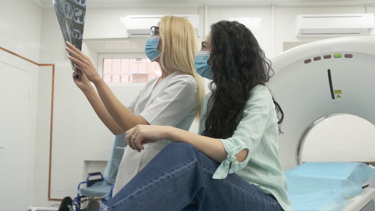 Woman doctor radiologist explains the results of CT scanning for young female patient, showing the snapshot with images, observing and analyzing CT scan in modern clinic beside modern CT scanner