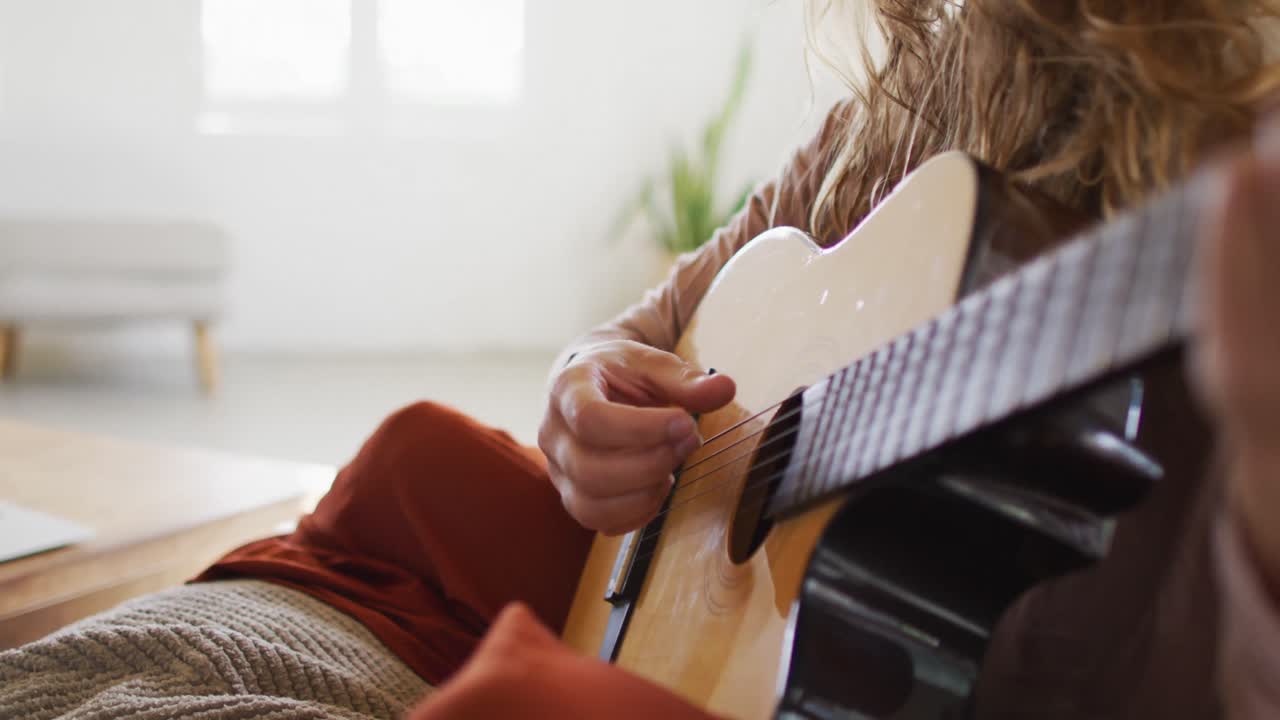 sección media de una mujer caucásica tocando guitarra acústica, sentada en la sala de estar de una cabaña soleada