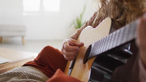 Midsection-of-caucasian-woman-playing-acoustic-guitar,-sitting-in-sunny-cottage-living-room