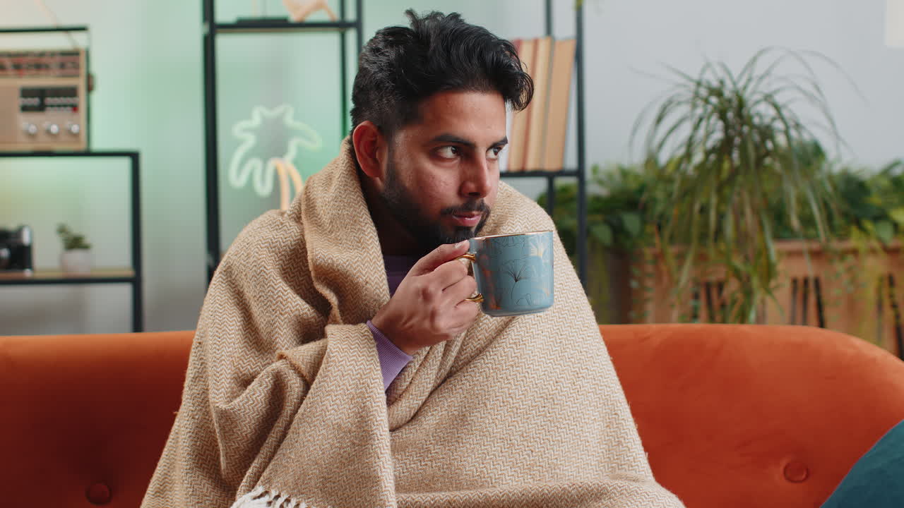 Uomo indiano malato avvolto in un plaid che trema dal freddo mentre beve tè caldo in un appartamento non riscaldato a causa dei debiti