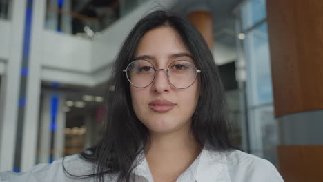 young woman in white shirt with long dark hair and glasses turns with a warm smile, standing near wooden and glass interior in modern space with blurred blinking blue light in background