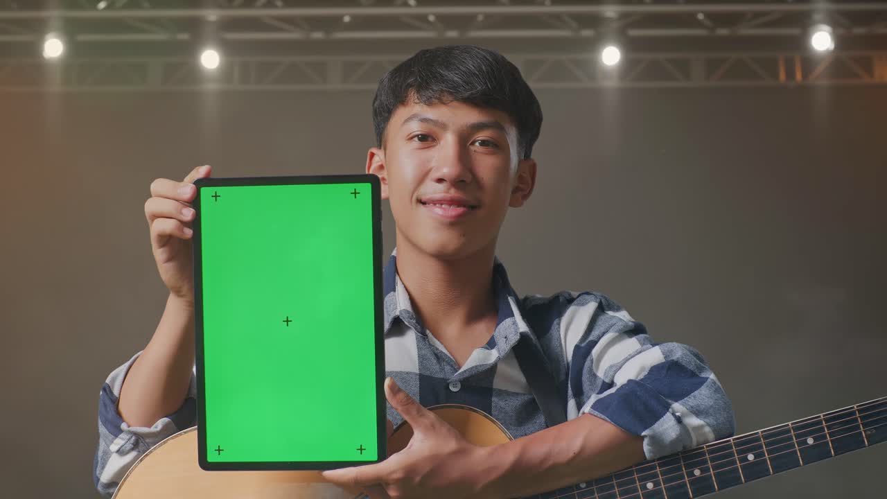무대에 서 있는 동안 기타를 웃고 녹색 화면 태블릿을 보여주는 아시아 십대의 클로즈업