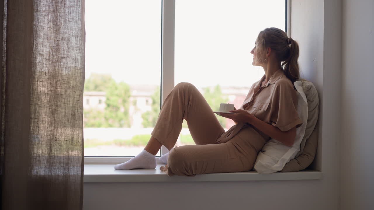 Una donna soddisfatta si rilassa con una tazza di tè sul davanzale della finestra.