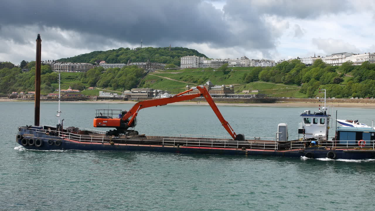 A barge carrying an orange excavator floats in Scarborough, North Yorkshire, England, with green hills and seaside architecture rising in the distance