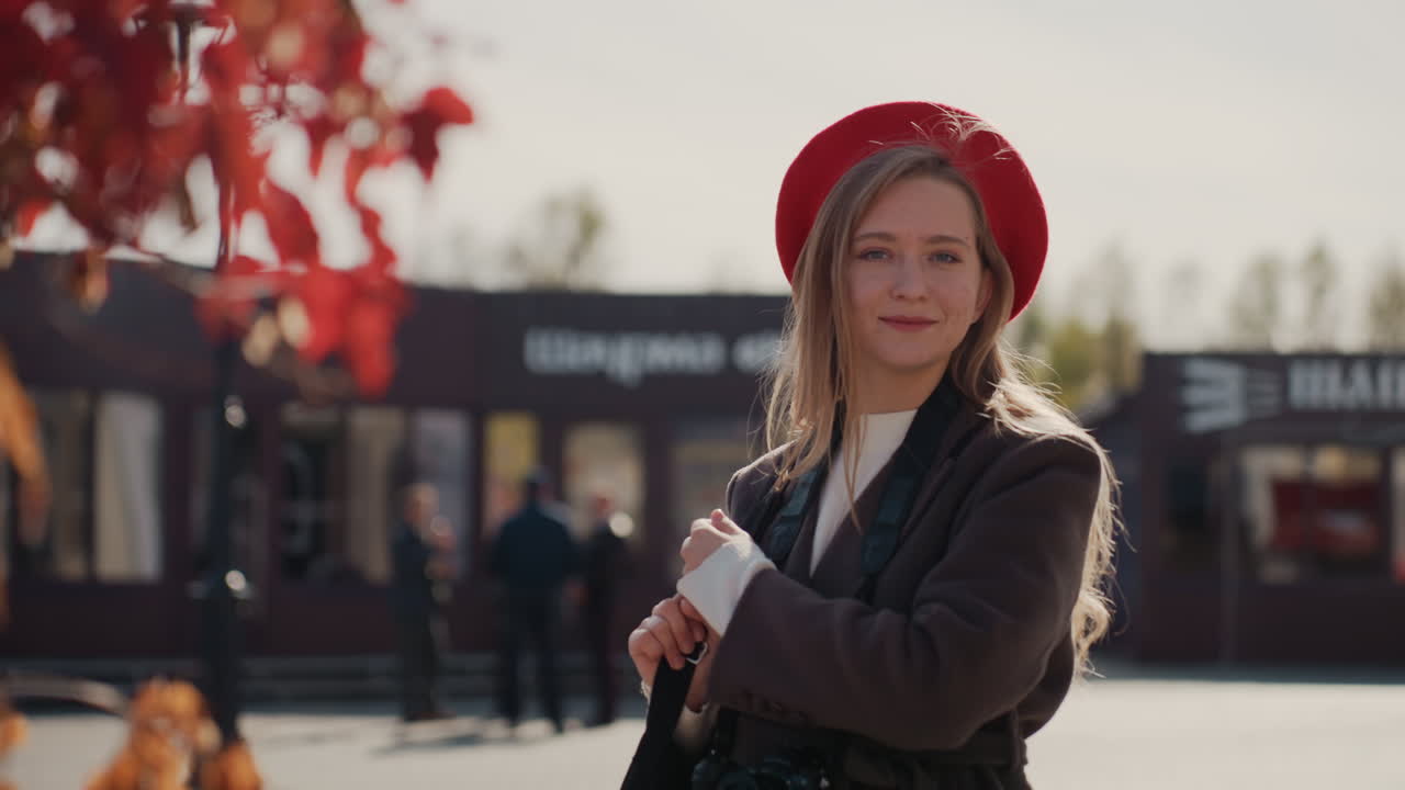 Ritratto ravvicinato di una donna caucasica sicura di sé con basco, sotto una calda luce autunnale. Foglie rosse le incorniciano il viso, tiene una tazza da asporto, indossa un cappotto alla moda, sfoggia un sorriso sottile e uno sguardo diretto, con obiettivo cinematografico, bokeh morbido, in contesto urbano.