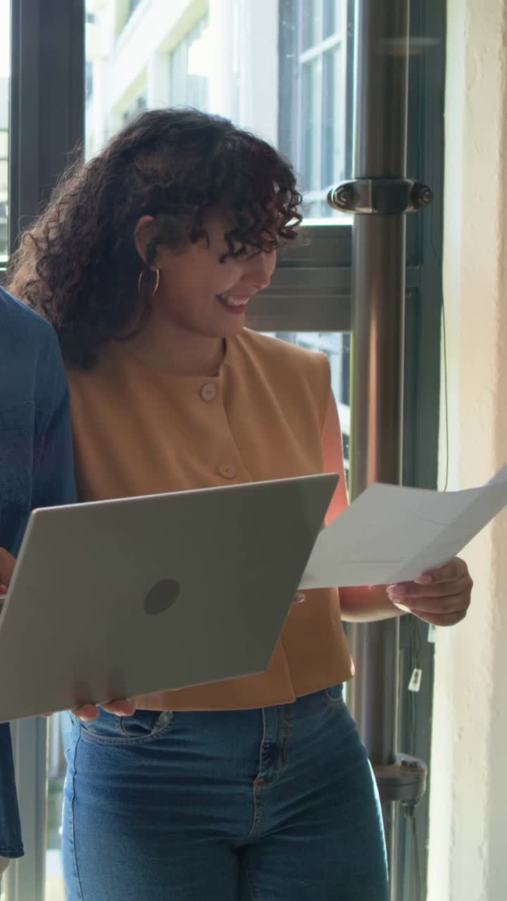 Vídeo vertical: Mulher lendo folha, com colete bege, notebook prateado e papéis junto à janela, revisando