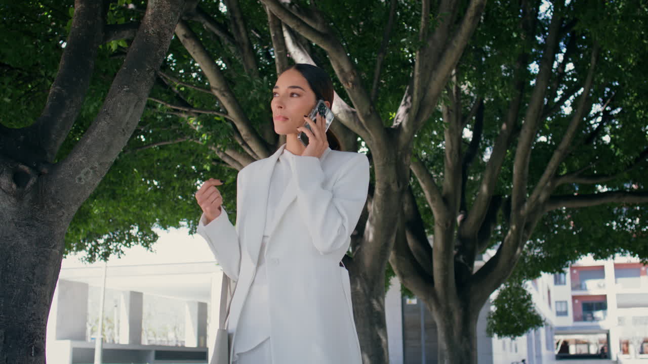 부유한 여성이 스마트폰으로 전화하고 우아한 색 슈트를 입고 공원 골목길을 고 있습니다.