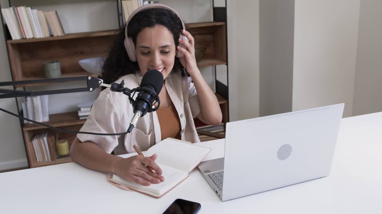 Mujer de ascendencia africana inclinada hacia el micrófono en el escritorio hablando, tomando notas, ajustando auriculares rosas y grabando