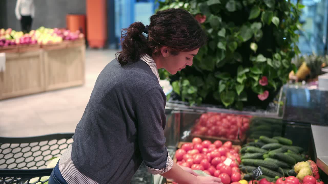 Young beautiful brunette girl in her 20's picking out tomatoes into a plastic bag at the fruit and vegetable aisle in a grocery store