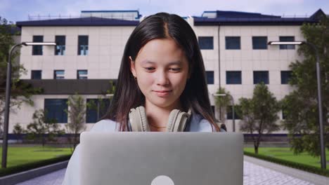 close up of asian teen girl student with a backpack smiling and typing on a laptop while standing in front of a school building