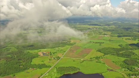 Aerial-drone-shot-through-clouds-over-green-rural-countryside-beside-a-lake-on-a-sunny-morning