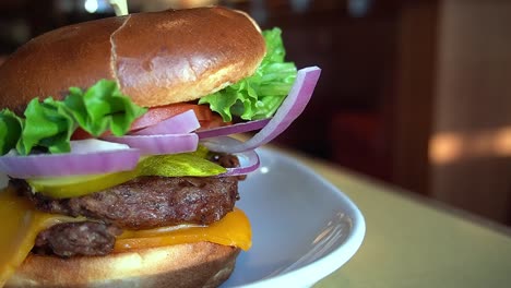 closeup rotating toable top double stack burger patties cheddar cheese pickles red onions tomatos lettuce in a kaiser pretzel bun on a white plate with mashed potato with a blurred bokeh background