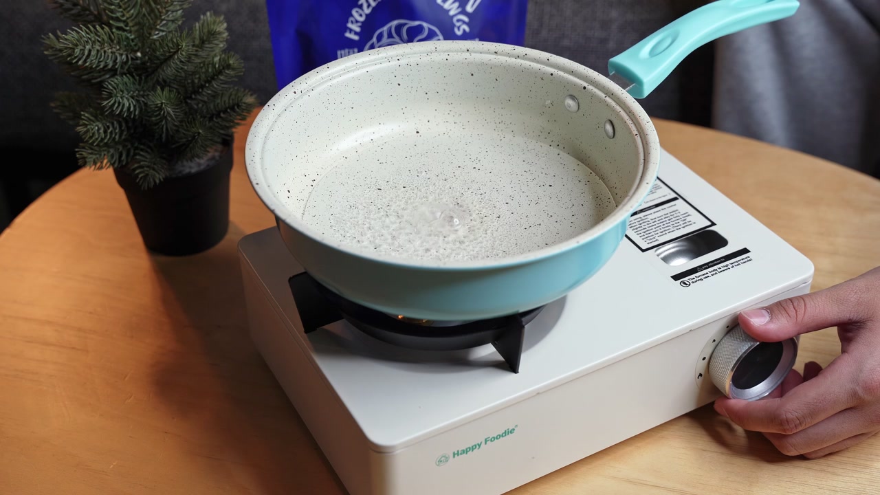Close-up of a person's hand turning the dial to ignite a white portable gas stove. A light blue speckled frying pan rests on the burner upon a round wooden table, getting ready to cook indoors.