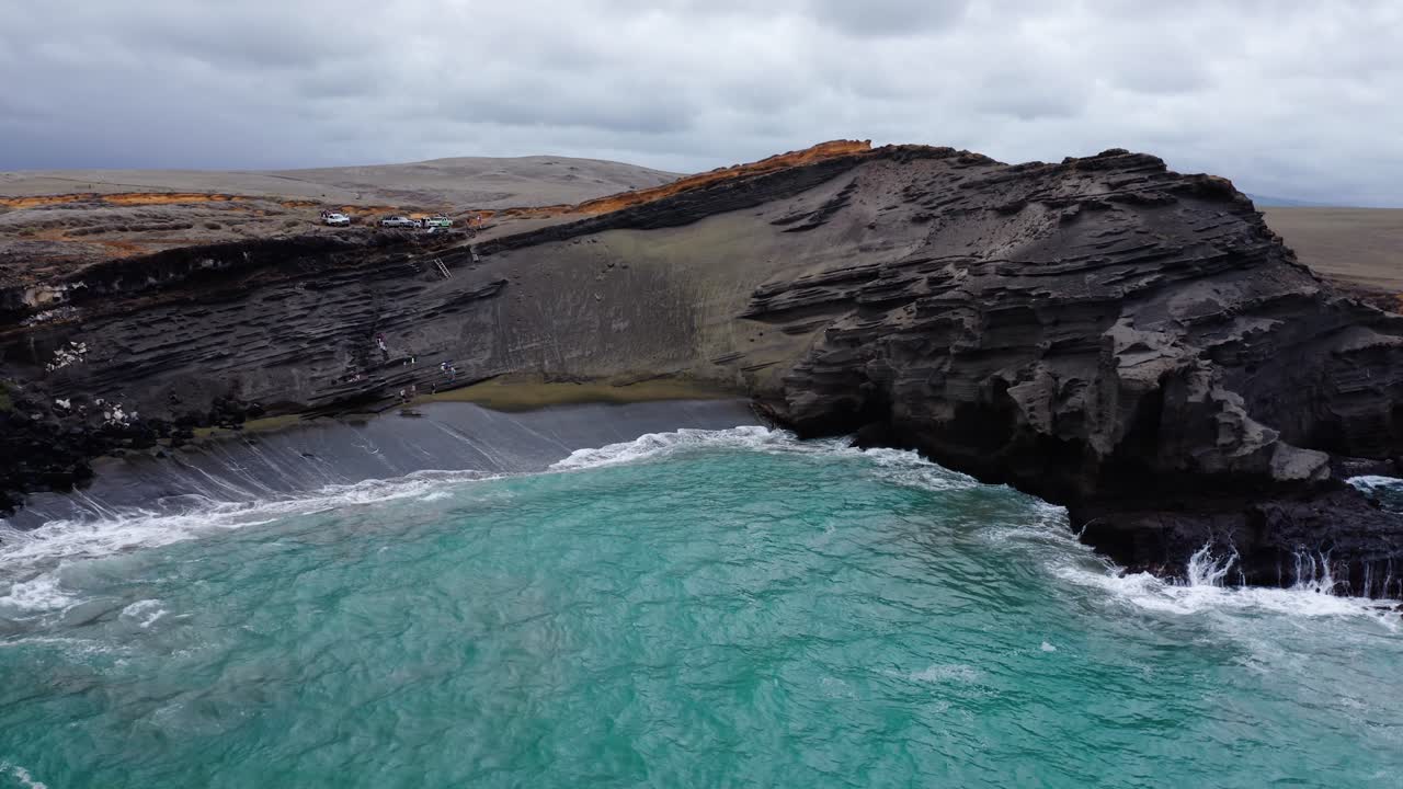 A vivid turquoise ocean meets a striking black sand beach framed by dramatic layered volcanic cliffs. The unique coastal landscape showcases raw natural beauty and rugged textures.