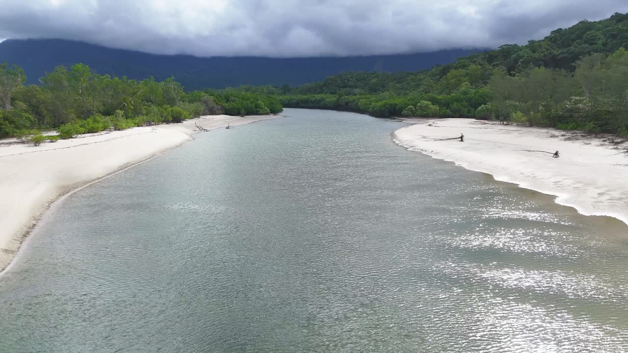 Drone glides above winding mangrove river, sandy banks, and rainforest under dramatic cloudy sky