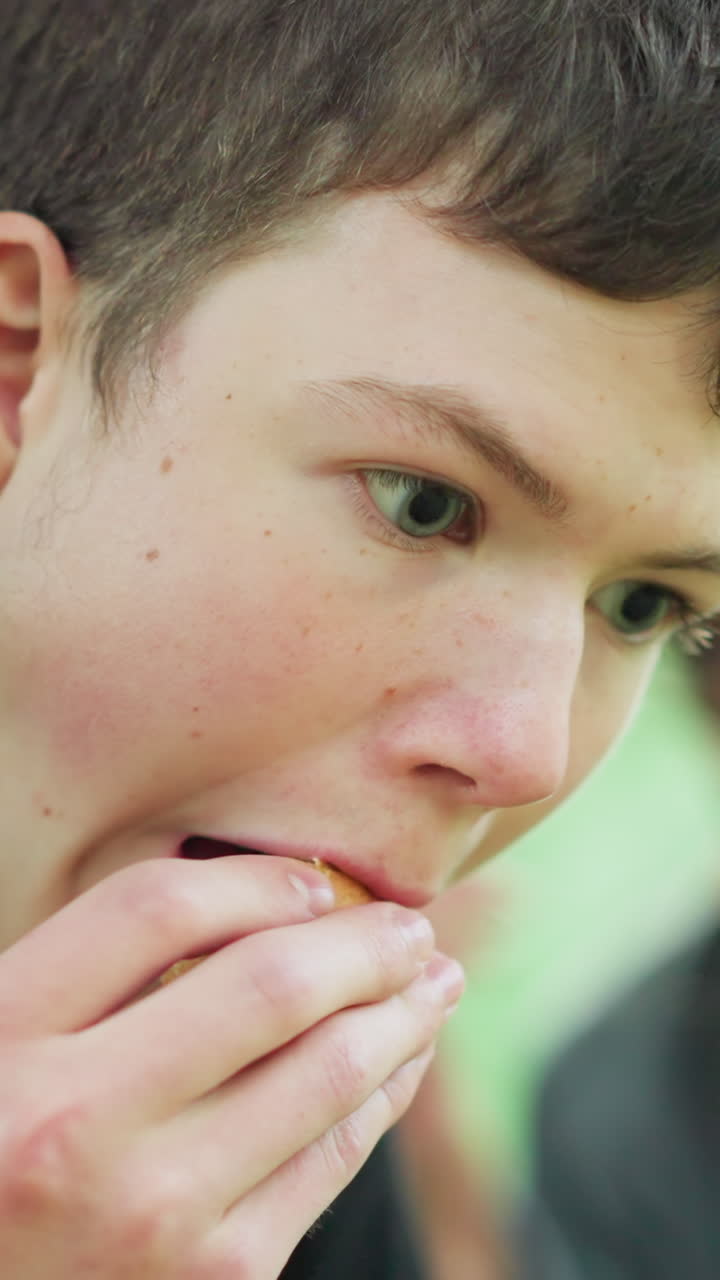 primer plano de un niño con una camiseta negra disfrutando de un bocadillo bajo un árbol, está centrado en su comida, mientras que en el fondo, una vista borrosa de otra persona también se ve comiendo bajo un árbol de hojas