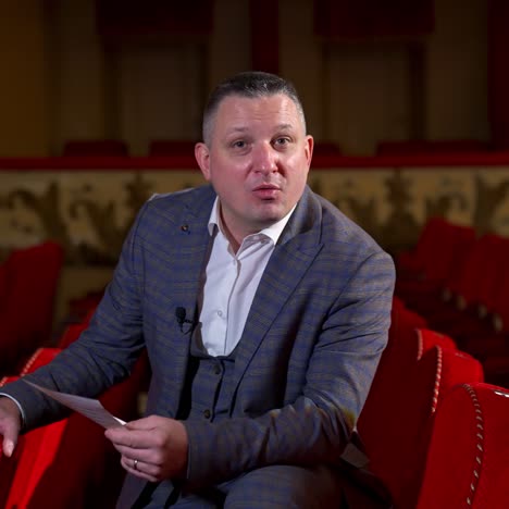 performer announces future theater season. actor is sitting in empty theater on red velvet chair with sheet of paper in hands