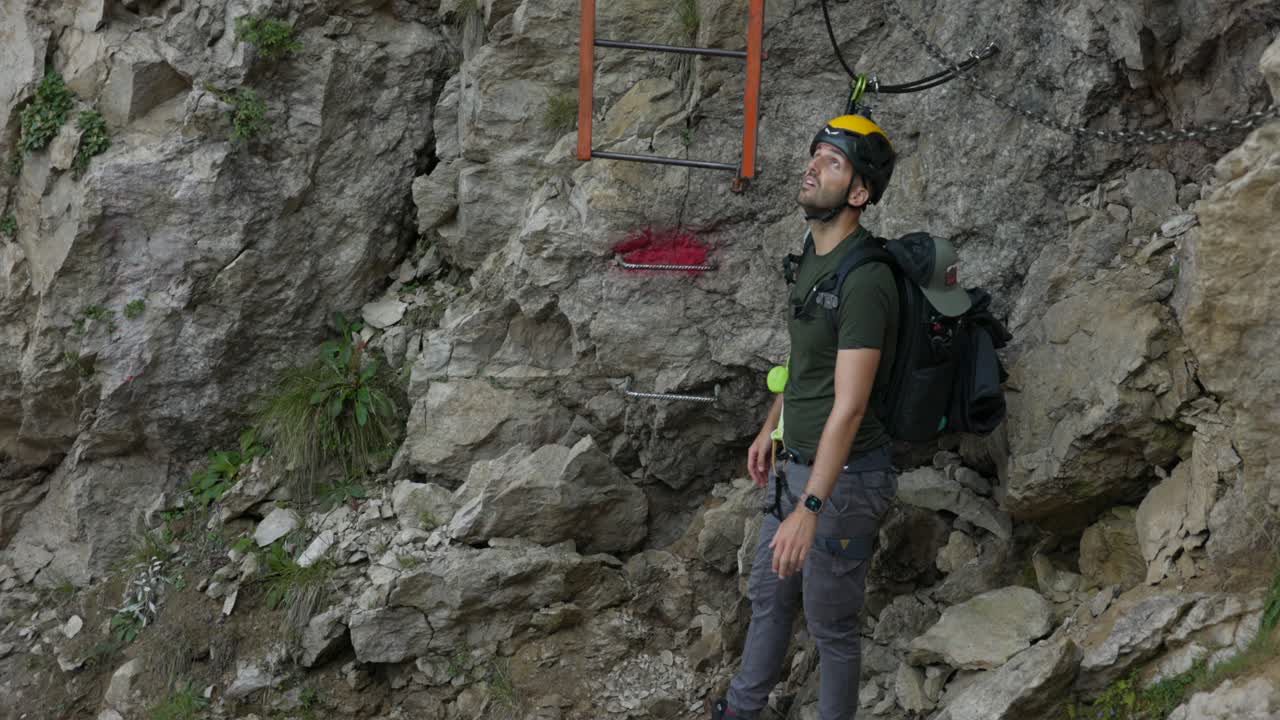 A man prepares to record himself with a drone on a rocky mountain trail, equipped with safety gear and a helmet. The scene highlights adventure, self-filming, and outdoor exploration
