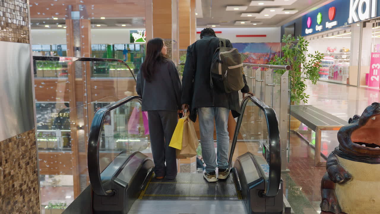 Rear view of couple holding colorful shopping bags stepping onto descending escalator inside modern shopping center with toy store, decorations, and open retail layout with polished floor reflections