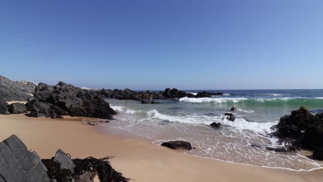 Man,-dog-and-woman-walking-in-beautiful-wavy-beach-with-big-rock-formations