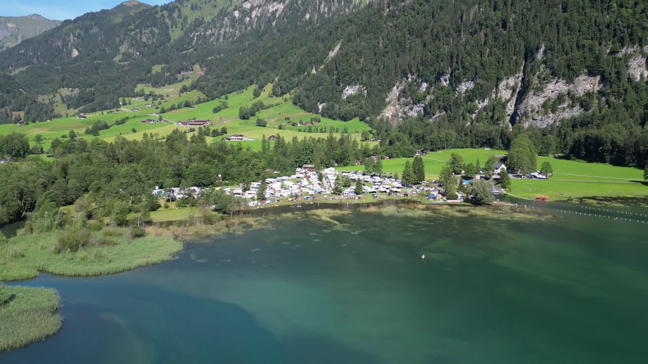 movimiento en órbita alrededor de un área de campamento donde las furgonetas blancas están estacionados con sus familias para unas vacaciones escapar aventura en la naturaleza bajo la colina masiva grandes montañas aguas puras azul vívido cinematográfico