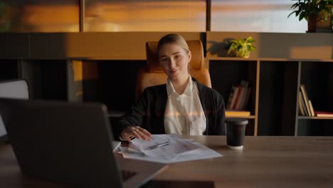 Portrait-of-a-confident-blonde-girl-in-a-business-uniform-who-sits-at-a-wooden-table-in-the-office-in-front-of-important-papers-and-a-laptop-in-a-sunny-office