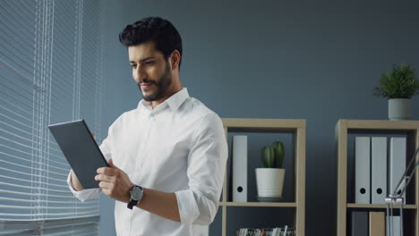 Smiled-Good-Looking-Arabian-Businessman-Standing-In-Comfortable-Office-And-Using-Tablet,-Then-Looking-Out-The-Window