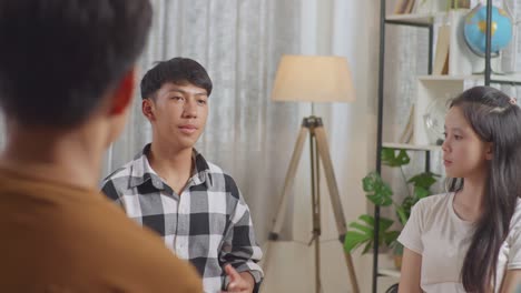 asian teen group sitting in chairs forming a circle discussing at home. close up of a boy in plaid shirt introducing himself