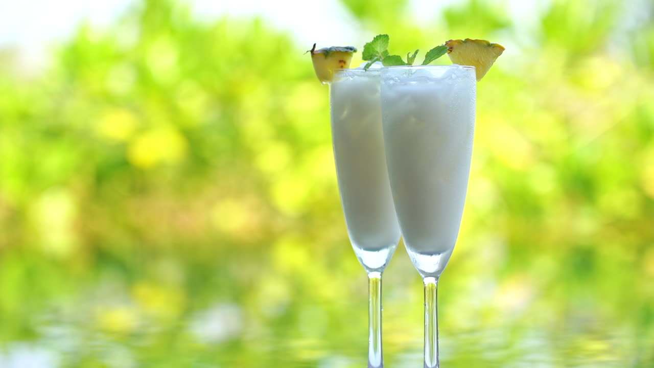 Static closeup shot of two frozen margarita glasses with a pool and trees blurred in the background