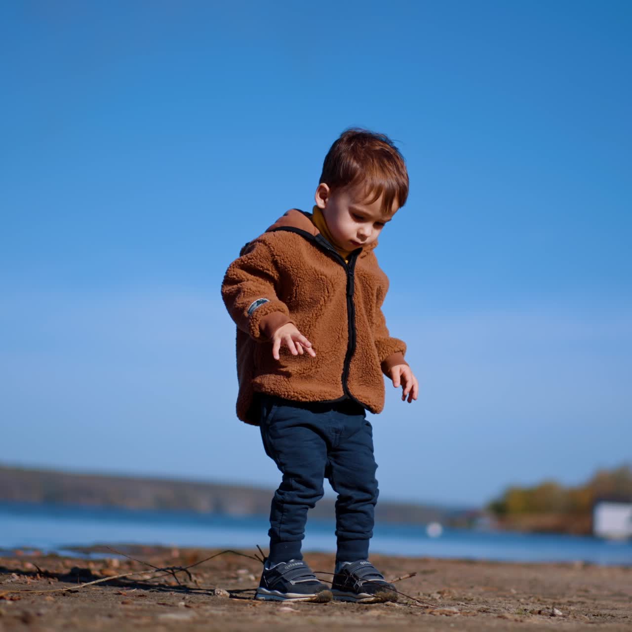 Cute kid walks by the waterfront looking for the stones. Baby boy touches a twig and then continues to go. Low angle view