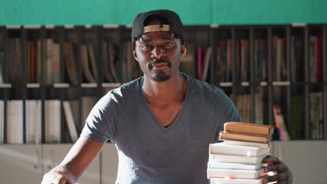 Man wearing cap sits at library table with stack of books, slowly raising head with eyes closed and thoughtful expression, teal wall and organized shelves behind him bathed in warm sunlight