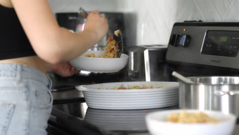 Caucasian-lady-scooping-a-serving-of-pasta-onto-a-white-bowl-and-placing-onto-a-counter