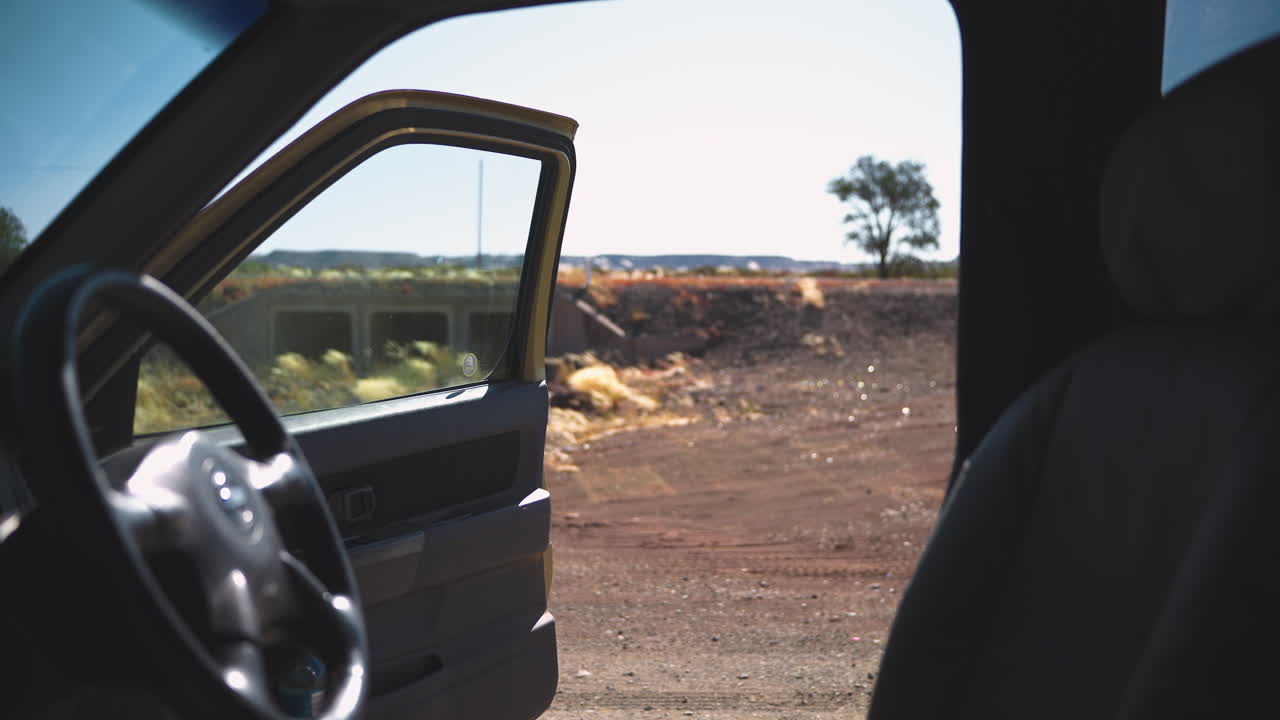 Static shot from inside a car parked in the Kalahari Desert, Namibia. The open door frames the arid landscape with scattered bushes under the bright midday sun
