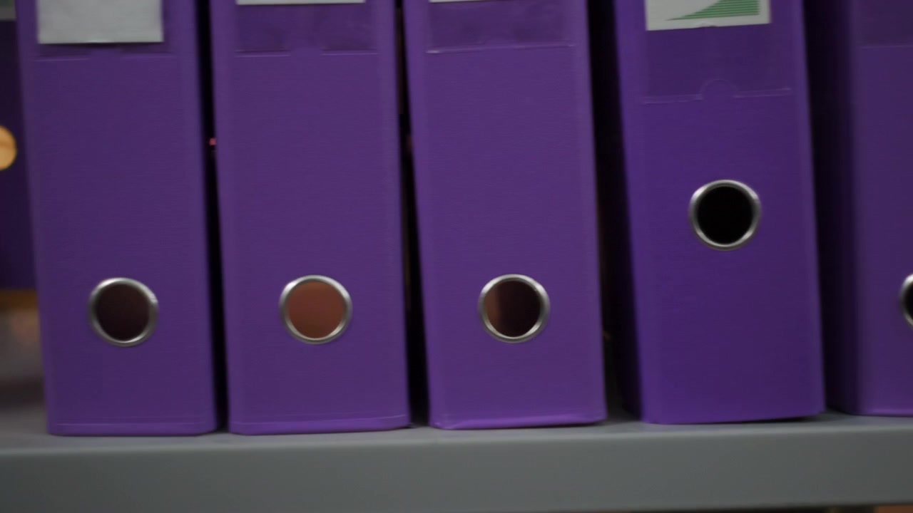Close-up side view of upright purple binders on office shelving, with one binder slightly pulled forward revealing narrow gap, circular finger holes, cardboard edges, tidy storage, muted light